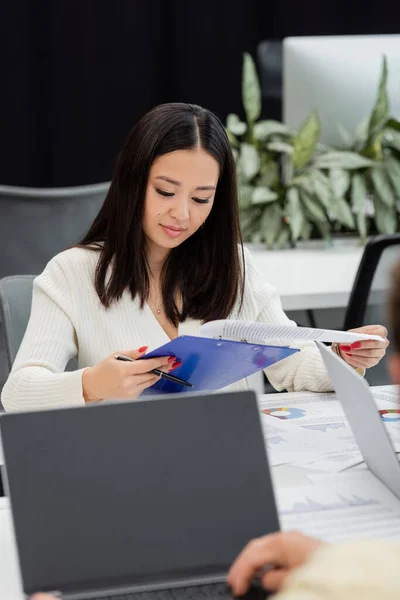 Giovane donna d'affari asiatica alla ricerca di documenti negli appunti mentre lavora in ufficio — Foto stock