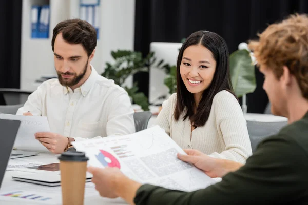 Cheerful asian businesswoman looking at camera near colleagues working with documents in office — Stock Photo