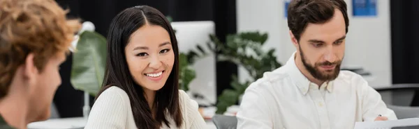 Brunette asian woman smiling at camera near businessmen in office, banner — Stock Photo