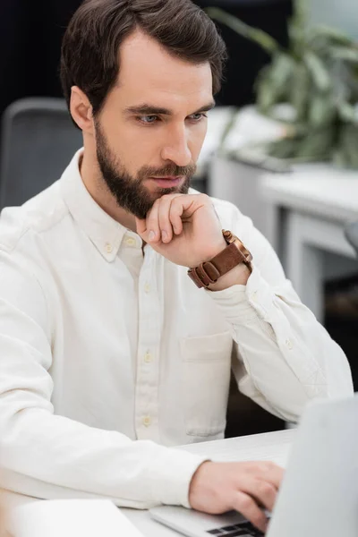 Вдумливий бородатий бізнесмен тримає руку біля обличчя і дивиться на розмитий ноутбук — стокове фото