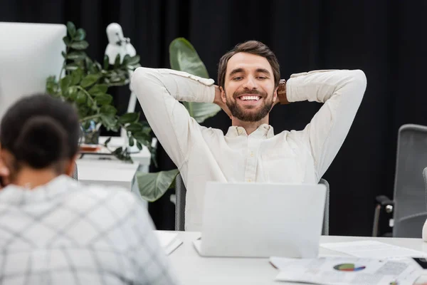 Happy bearded businessman sitting with hands behind back while looking at laptop near blurred multiracial woman — Stock Photo