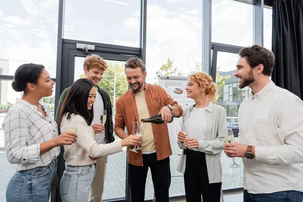 Cheerful man pouring champagne near multicultural friends during corporate party — Stock Photo