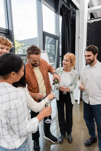 Businessman pouring champagne near cheerful interracial colleagues at corporate party — Stock Photo