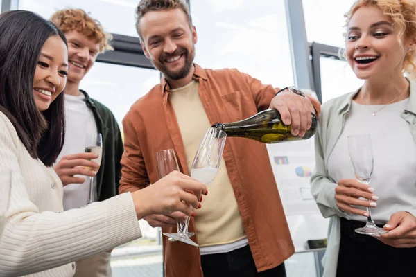 Cheerful bearded businessman pouring champagne at corporate party with excited interracial colleagues — Stock Photo