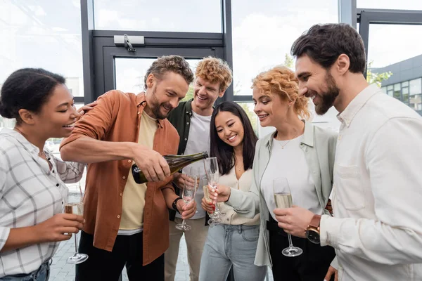 Bearded man pouring champagne during corporate party with cheerful multiethnic colleagues — Stock Photo