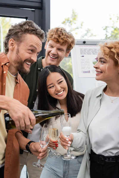 Hombre emocionado verter champán cerca de compañeros de negocios multiculturales felices durante la fiesta corporativa - foto de stock
