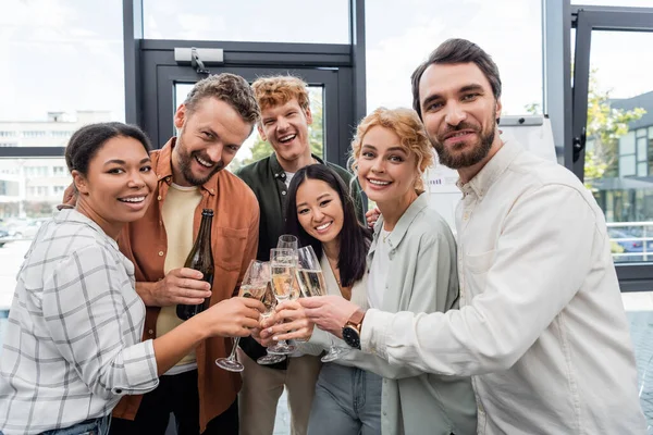 Colegas de negócios multiétnicos sorrindo para a câmera e batendo copos de champanhe na festa corporativa — Fotografia de Stock