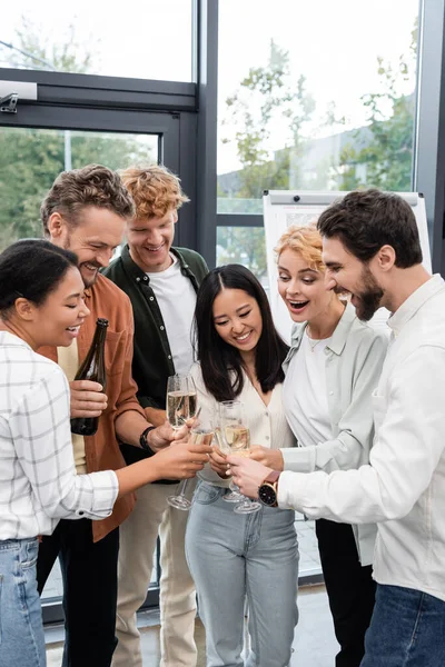 Gente d'affari multietnica felice che tiene occhiali vicino a collega con bottiglia di champagne in ufficio — Foto stock