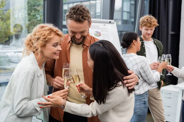Lächelnder Geschäftsmann umarmt interrassische Kollegen im Büro mit Champagner — Stockfoto