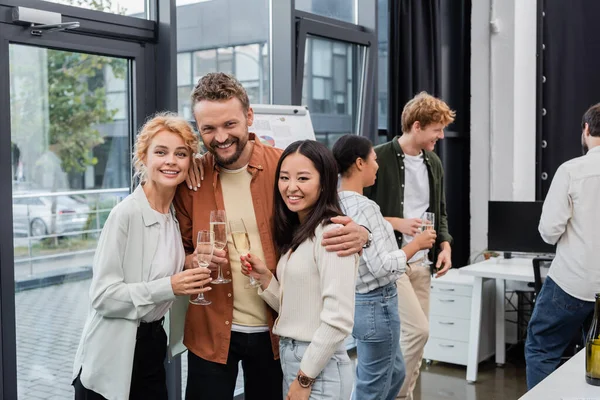 Empresario positivo abrazando a colegas multiétnicos con champán en la oficina — Stock Photo