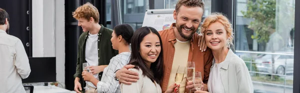 Interracial business people hugging and holding glasses of champagne in office, banner — Stock Photo