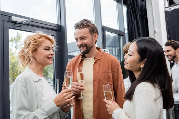 Smiling businesswoman talking to interracial colleagues with glasses of champagne in office — Stock Photo