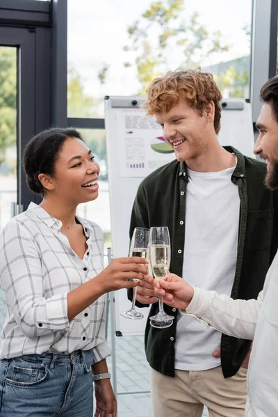 Cheerful multiracial businesswoman clinking champagne with colleagues in office — Stock Photo