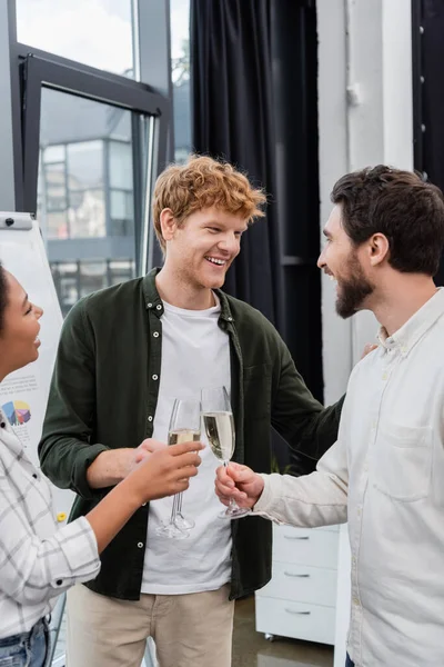 Smiling redhead businessman talking to interracial colleagues with champagne in office — Stock Photo