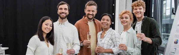 Мультикультурні бізнесмени тримають келихи шампанського і дивляться на камеру в офісі, банер — стокове фото