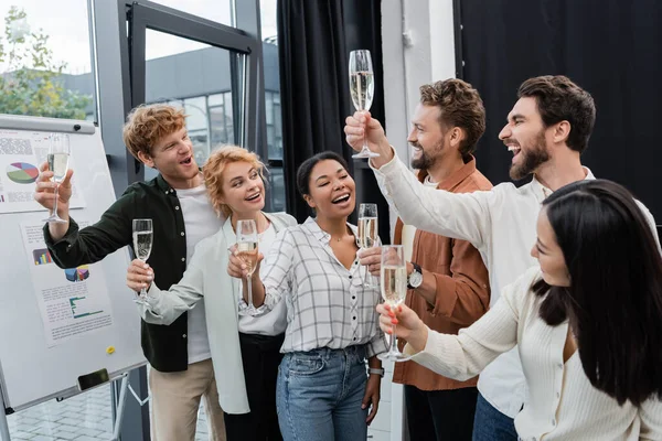 Gente de negocios interracial emocionada brindando y sosteniendo copas de champán cerca del rotafolio en la oficina - foto de stock