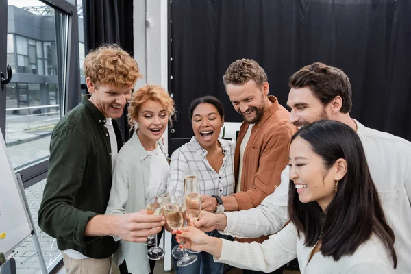 Empresários multiculturais entusiasmados olhando para copos de champanhe enquanto clica no escritório — Fotografia de Stock