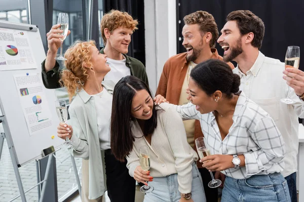 Cheerful multiethnic business people talking and holding glasses of champagne in office — Stock Photo