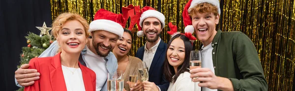 Des hommes d'affaires interraciaux joyeux dans des chapeaux de Père Noël tenant du champagne près de tinsel au bureau, bannière — Photo de stock