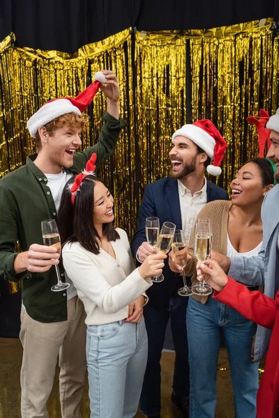 Positive interrassische Geschäftsleute blicken auf Kollegen mit Weihnachtsmütze und halten Champagner im Büro — Stockfoto