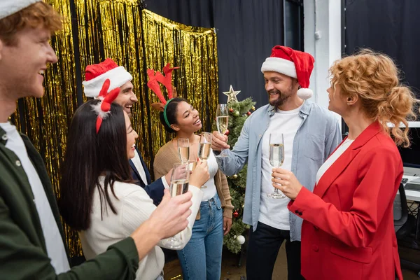 Smiling businessman in santa hat toasting and holding champagne near interracial colleagues in office — Stock Photo