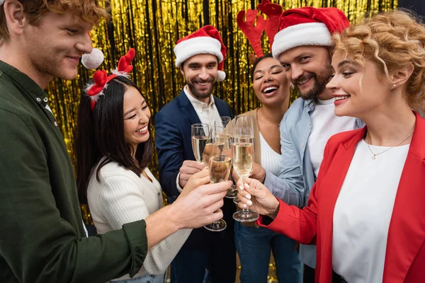 Sorridenti uomini d'affari multiculturali in cappelli di Babbo Natale clinking champagne vicino fili di lame sfocati in ufficio — Foto stock