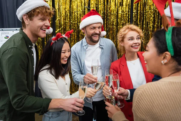 Glückliche multikulturelle Geschäftsleute in Weihnachtsmützen und Stirnbändern, die Champagner im Büro halten — Stockfoto