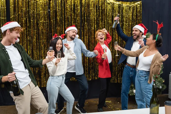 Positive multikulturelle Geschäftsleute mit Weihnachtsmannhüten, die Champagner in der Hand halten und im Büro tanzen — Stockfoto