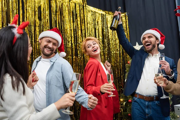 Aufgeregter Geschäftsmann mit Weihnachtsmann-Hut, der eine Flasche Champagner in der Nähe von interrassischen Kollegen hält, die im Büro tanzen — Stockfoto