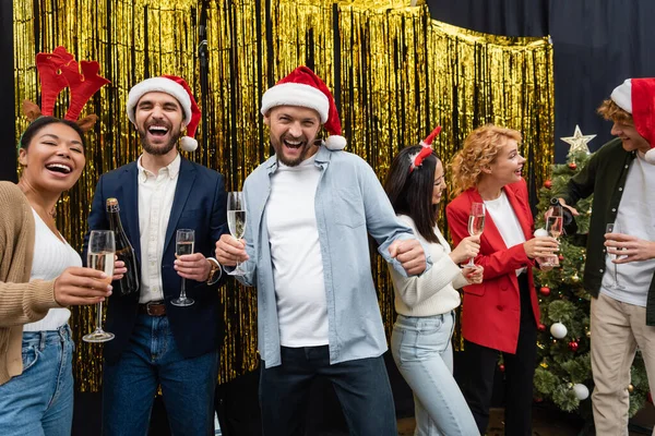 Empresários multiétnicos alegres em chapéus de Santa segurando champanhe perto de colegas e ouropel no escritório — Fotografia de Stock