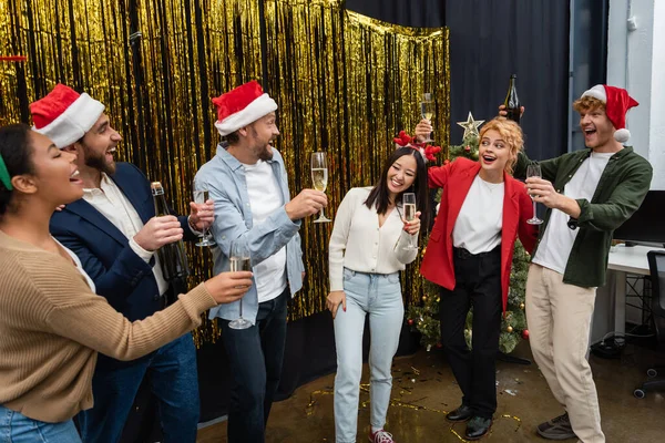 Positivi uomini d'affari multiculturali in cappelli di Babbo Natale in possesso di champagne vicino orpelli in ufficio — Foto stock