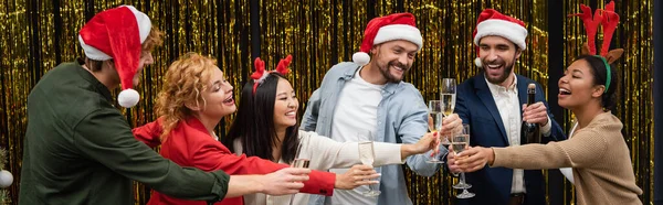 Positivi uomini d'affari multietnici in cappelli di Babbo Natale clinking champagne con colleghi vicino orpelli, banner — Foto stock