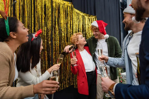 Улыбающийся бизнесмен наливает шампанское рядом с межрасовыми коллегами в очках во время рождественской вечеринки в офисе — стоковое фото