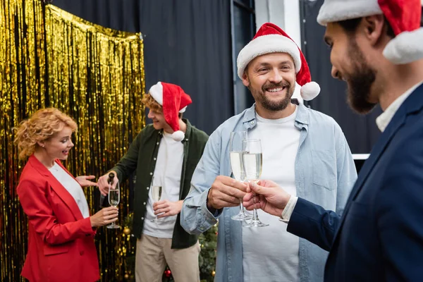 Улыбающиеся бизнесмены в шляпах Санты держат шампанское рядом с размытыми коллегами во время рождественской вечеринки в офисе — стоковое фото