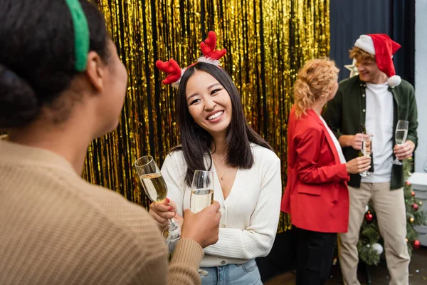 Улыбающаяся азиатская бизнесвумен, держащая шампанское и разговаривающая с коллегой по многонациональности во время рождественской вечеринки в офисе — стоковое фото