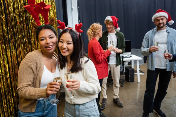 Позитивные межрасовые деловые женщины в рождественских повязках, держащие шампанское возле мишуры в офисе — стоковое фото