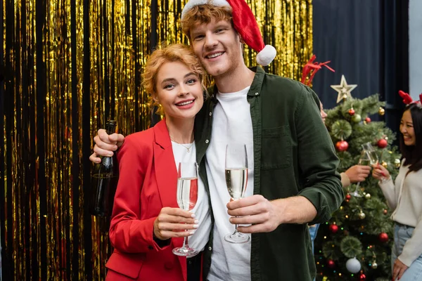 Pessoas de negócios sorridentes com champanhe olhando para a câmera perto de ouropel durante a festa de Natal no escritório — Fotografia de Stock