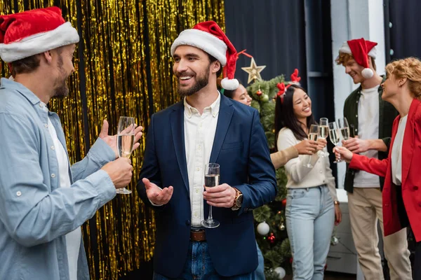 Lächelnder Geschäftsmann mit Weihnachtsmannmütze hält Champagner in der Hand und spricht mit Amtskollegin — Stockfoto