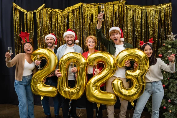 Pessoas de negócios inter-raciais alegres segurando champanhe e balões em forma de 2023 no escritório — Fotografia de Stock
