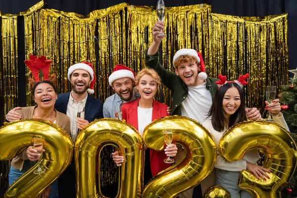 Positive multiethnic business people with champagne looking at camera near balloons in shape of 2023 sign in office — Stock Photo