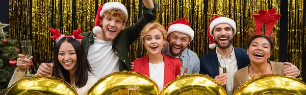 Веселые многонациональные бизнесмены, держащие бокалы шампанского возле воздушных шаров во время новогодней вечеринки в офисе, баннер — стоковое фото