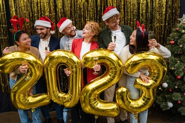 Positive multicultural business people in santa hats holding champagne near balloons in shape of 2023 in office — Stock Photo