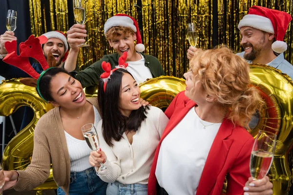 Mujeres de negocios multiétnicas sonrientes sosteniendo champán cerca de globos y hombres de negocios durante la fiesta de Navidad en la oficina - foto de stock