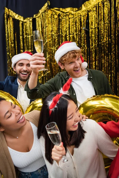 Веселые многонациональные бизнесмены, держащие шампанское возле воздушных шаров и мишуры во время новогодней вечеринки в офисе — стоковое фото