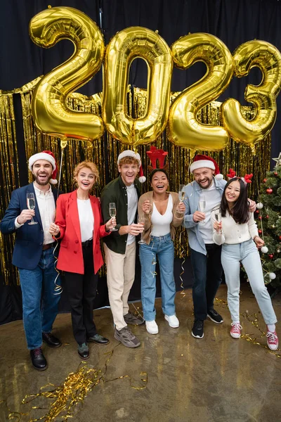 Empresários multiculturais positivos em chapéus de Papai Noel segurando champanhe perto de balões em 2023 forma no escritório — Fotografia de Stock