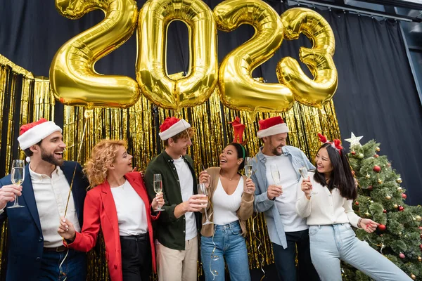 Smiling multiethnic business people holding champagne and balloons in shape of 2023 signs in office — Stock Photo