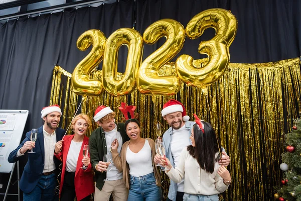 Multiethnic business people looking at asian colleague with champagne near balloons in shape of 2023 signs in office — Stock Photo