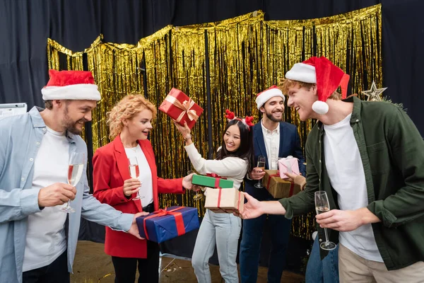 Sorridenti uomini d'affari multietnici in cappelli di Babbo Natale in possesso di champagne e regali in ufficio — Foto stock