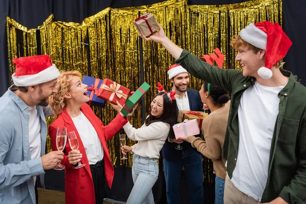 Багатоетнічні ділові люди в капелюхах Санти тримають подарунки та шампанське під час різдвяної корпоративної вечірки в офісі — стокове фото