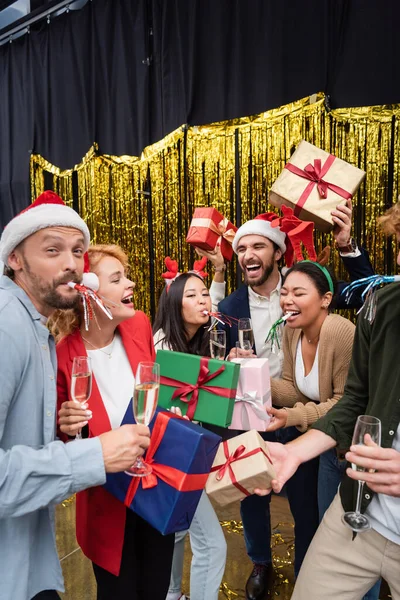 Веселі міжрасові ділові люди з партійними рогами тримають подарунки та шампанське в офісі — стокове фото